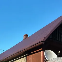 Ciemnobrązowy dach z blachy falistej, ceglany komin, rynna i antena satelitarna na tle błękitnego nieba.