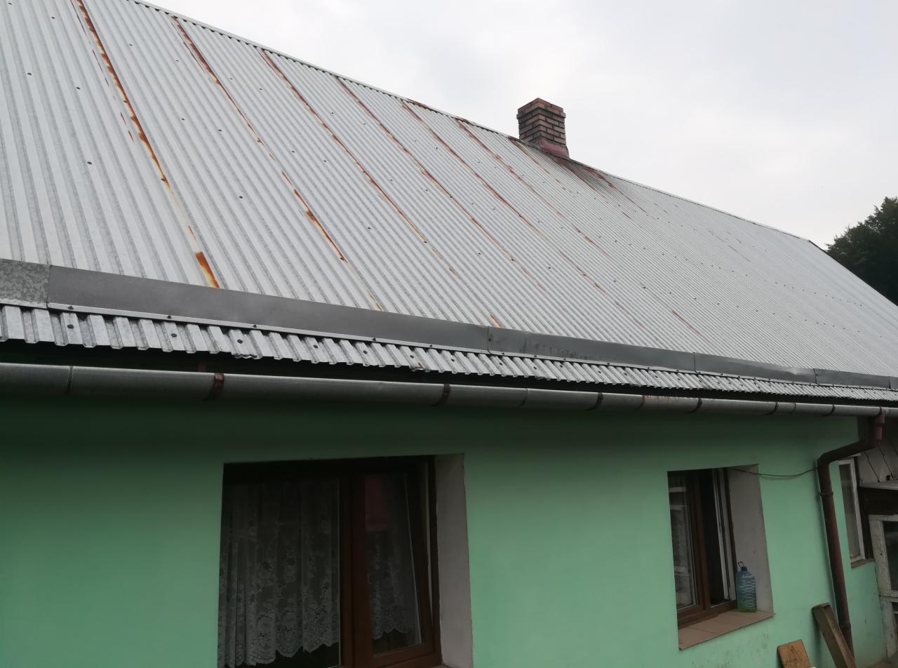 Dom z zieloną elewacją i blachodachówką z widocznymi śladami korozji, rynny zamontowane, widoczny komin z cegły.