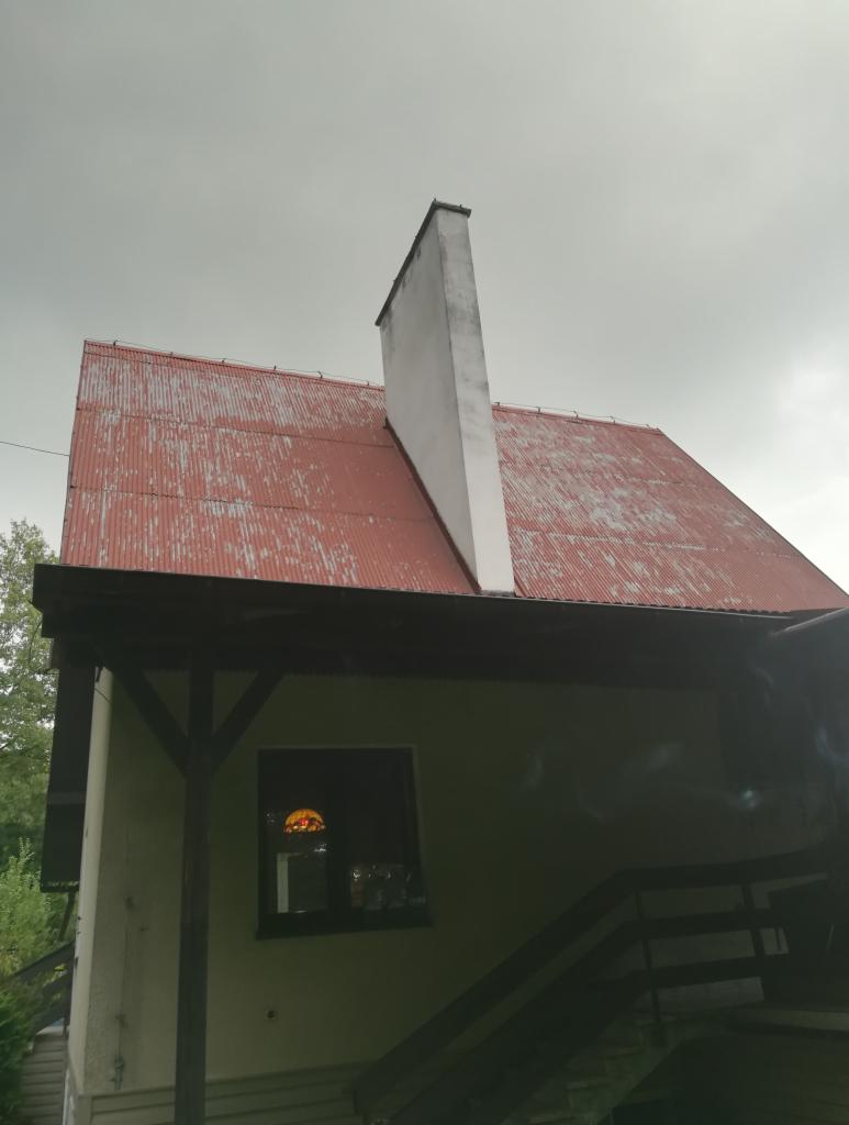 Czerwony, blaszany dach z białym kominem na tle pochmurnego nieba, widok z dołu na dom z werandą i schodami.