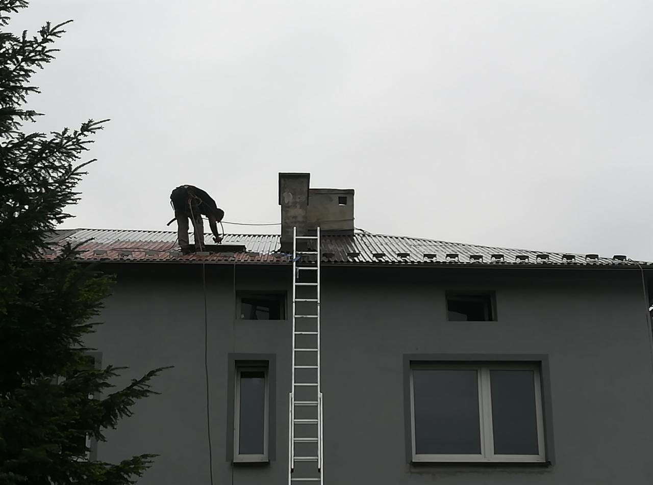 Pracownik w uprzęży na dachu budynku mieszkalnego, zabezpieczony liną, obok komina i drabiny, podczas prac dekarskich.