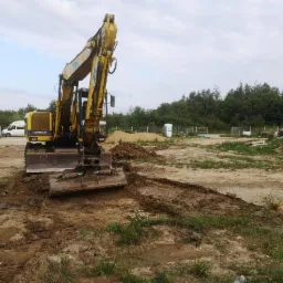 Żółta koparka Caterpillar na placu budowy, widoczny świeżo wyrównany teren z błotem, w tle biały bus, przenośna toaleta i czarny samochód osobowy.