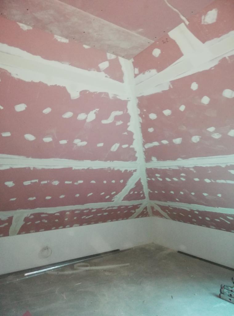 Różowa ściana z płyt kartonowo-gipsowych ze szpachlowanymi łączeniami i wkrętami, widok na narożnik pomieszczenia w trakcie prac wykończeniowych.