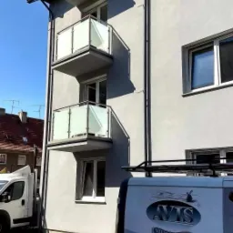 Trzy balkony w bloku mieszkalnym z nowoczesnymi balustradami ze szkła mlecznego i metalu, widoczne z poziomu ulicy obok zaparkowanego samochodu dostawczego z logo firmy.