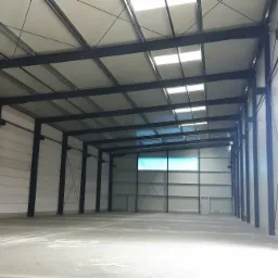 Wnętrze nowej hali magazynowej z blachy trapezowej i stalowej konstrukcji, widoczne świetliki dachowe i betonowa posadzka.