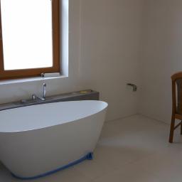 SwiFTbud - Nowoczesna, wolnostojąca wanna owalna z baterią podtynkową, osadzona na podwyższeniu, zabezpieczona niebieską folią ochronną, w minimalistycznej łazience z drewnianym oknem i krzesłem obok kabiny...