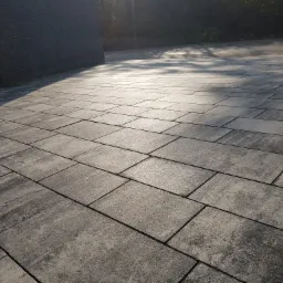 Ułożona, prostokątna, szara kostka na podjeździe, z widocznym cieniem drzew i fragmentem muru z kostki po lewej stronie.