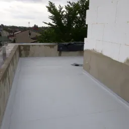 Płaski dach pokryty jasnoszarą membraną, widoczne obróbki blacharskie przy ścianie i attyce, w tle zabudowa jednorodzinna i zieleń.