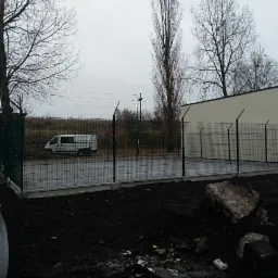 Zabezpieczony teren zieloną siatką ogrodzeniową z ostrymi zakończeniami, widoczny szary betonowy fundament, obok zaparkowany srebrny samochód, w tle budynek i drzewa bez liści.