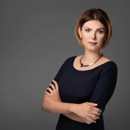 Portret kobiety z brązowymi włosami do ramion, ubranej w granatową sukienkę i srebrny naszyjnik, pozującej na szarym tle ze skrzyżowanymi ramionami.