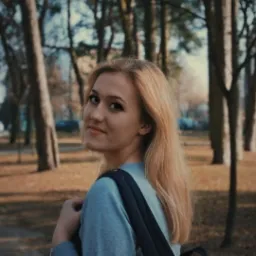 Młoda kobieta z blond włosami i plecakiem, odwrócona w stronę kamery, uśmiecha się na tle parku z drzewami.