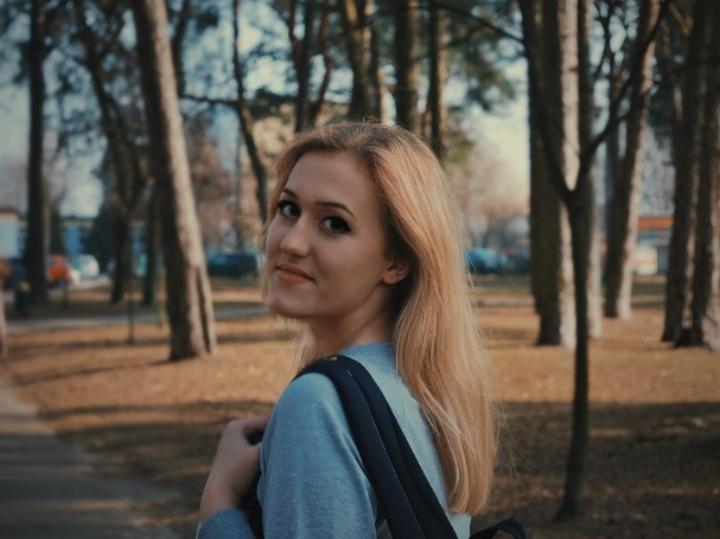 Młoda kobieta z blond włosami i plecakiem, odwrócona w stronę kamery, uśmiecha się na tle parku z drzewami.