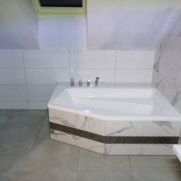 MIKA Michał Białek - Biała wanna narożna z marmurowym wykończeniem i mozaikowym pasem w łazience na poddaszu, widoczna umywalka i bidet, szare płytki podłogowe.