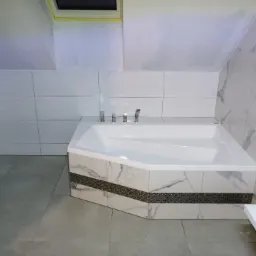 Biała wanna narożna z marmurowym wykończeniem i mozaikowym pasem w łazience na poddaszu, widoczna umywalka i bidet, szare płytki podłogowe.