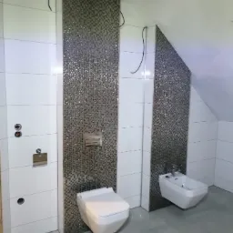 Nowoczesna łazienka w trakcie wykończenia, z białą armaturą i pionowymi pasami dekoracyjnej mozaiki, widoczne wystające kable i niezamaskowane otwory w ścianie.