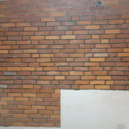 Ściana z cegieł klinkierowych w odcieniach czerwieni i brązu, częściowo wykończona, z widocznym otworem na instalację i fragmentem nieotynkowanej ściany poniżej.