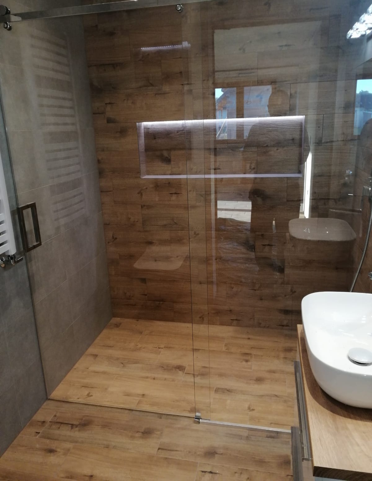 Nowoczesna kabina prysznicowa typu walk-in z drewnopodobnymi płytkami na ścianach i podłodze, wbudowana półka z oświetleniem LED, umywalka nablatowa.