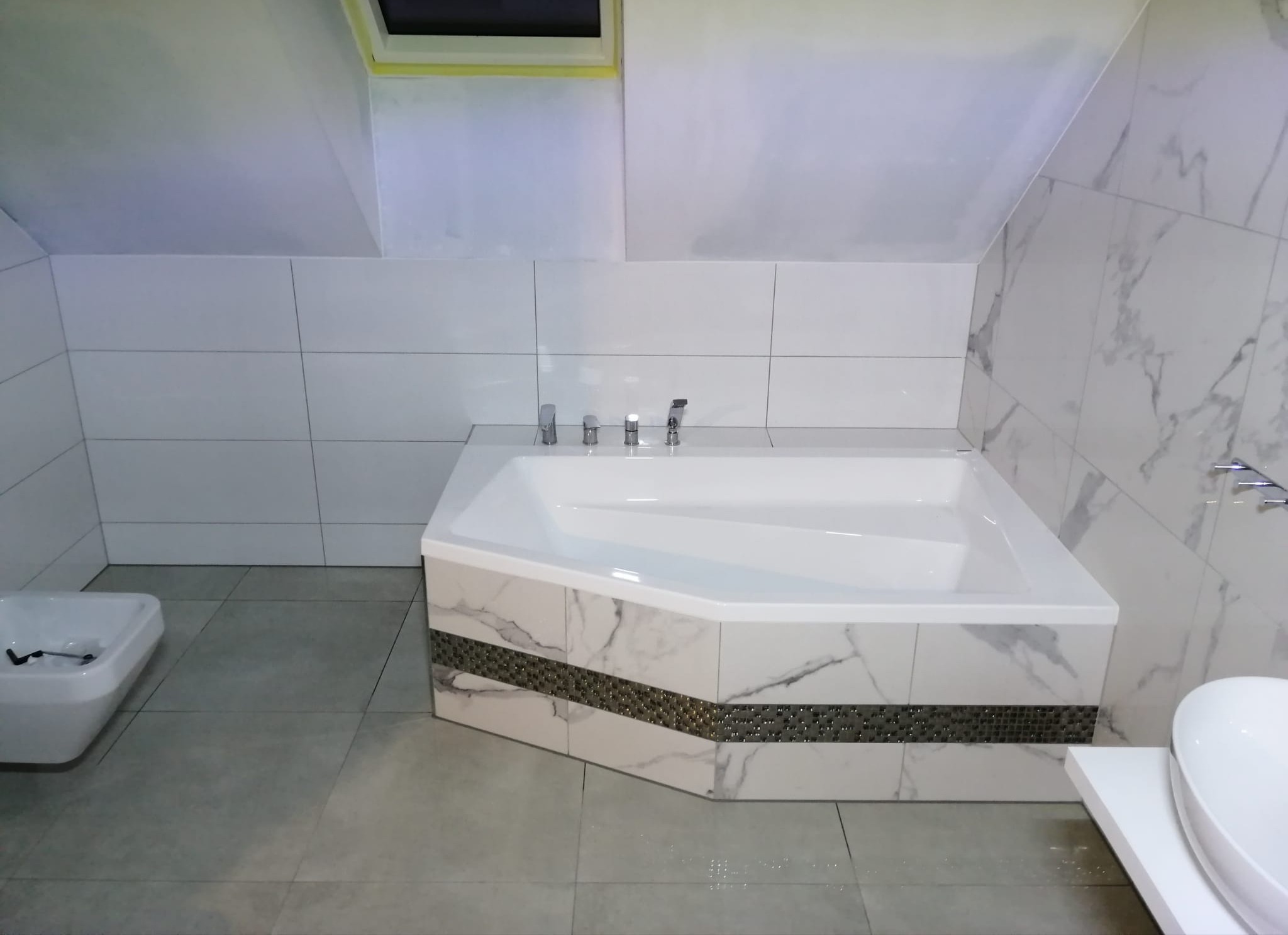 Biała wanna narożna z marmurowym wykończeniem i mozaikowym pasem w łazience na poddaszu, widoczna umywalka i bidet, szare płytki podłogowe.