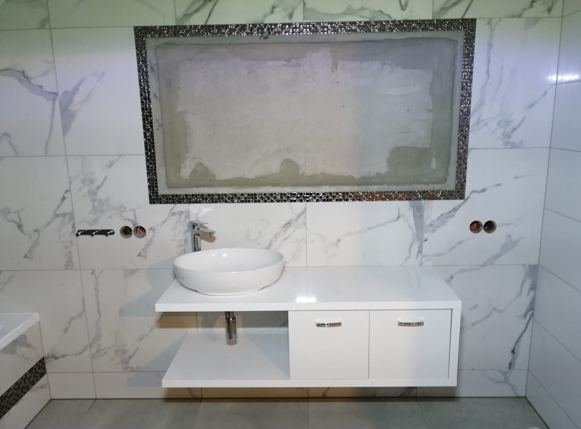 Łazienka w trakcie wykańczania: umywalka nablatowa na białej szafce, ściany wyłożone płytkami imitującymi marmur, wnęka z mozaiką przygotowana do dalszych prac.