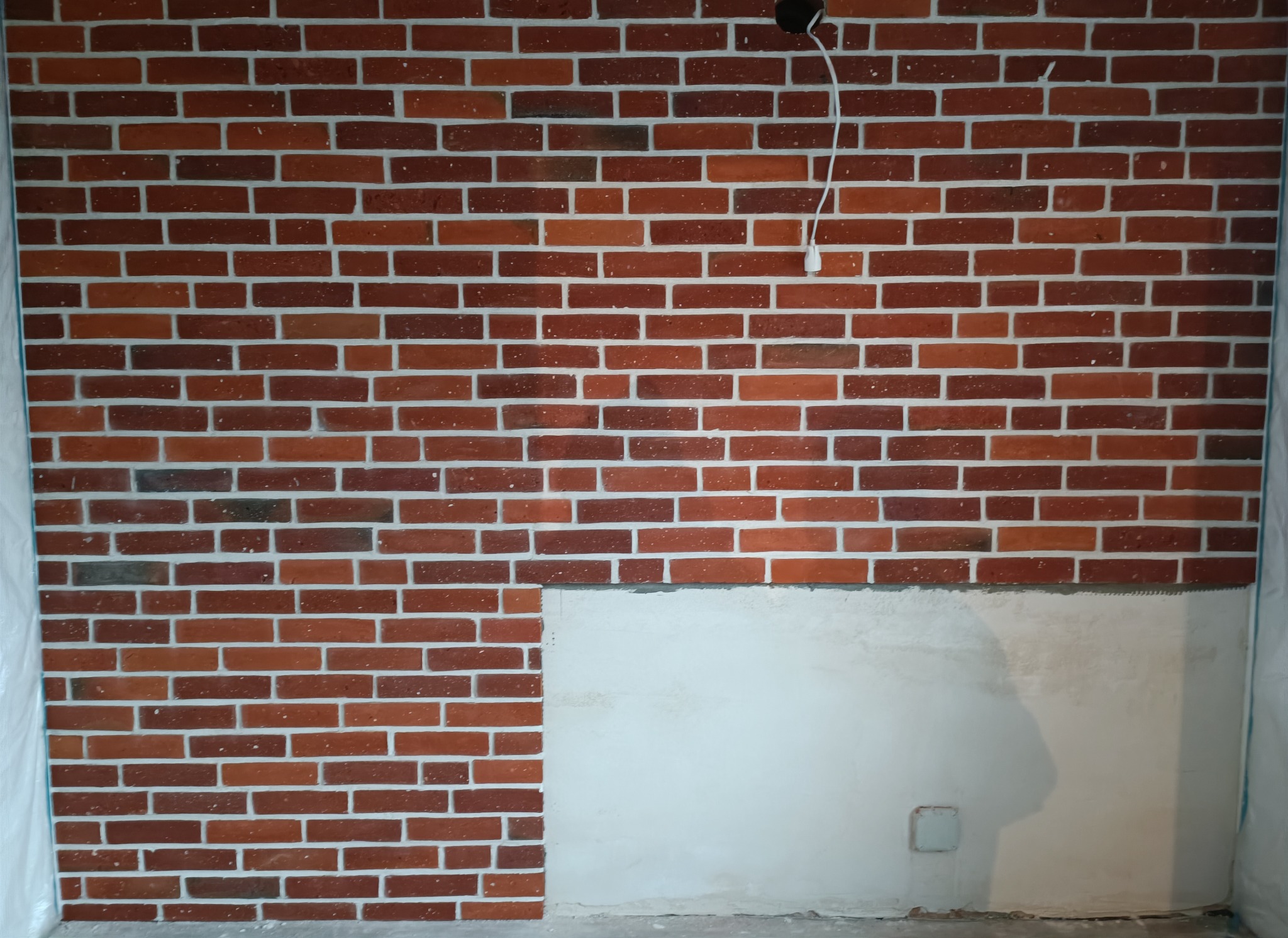 Ściana z czerwonej cegły klinkierowej z jasną fugą, częściowo wykończona, widoczne kable oświetleniowe i fragment ściany przygotowanej pod dalsze prace wykończeniowe.