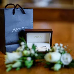Dwa srebrne obrączki w drewnianym pudełku marki Apart, otoczone białymi kwiatami, obok firmowa torba na drewnianym stole.
