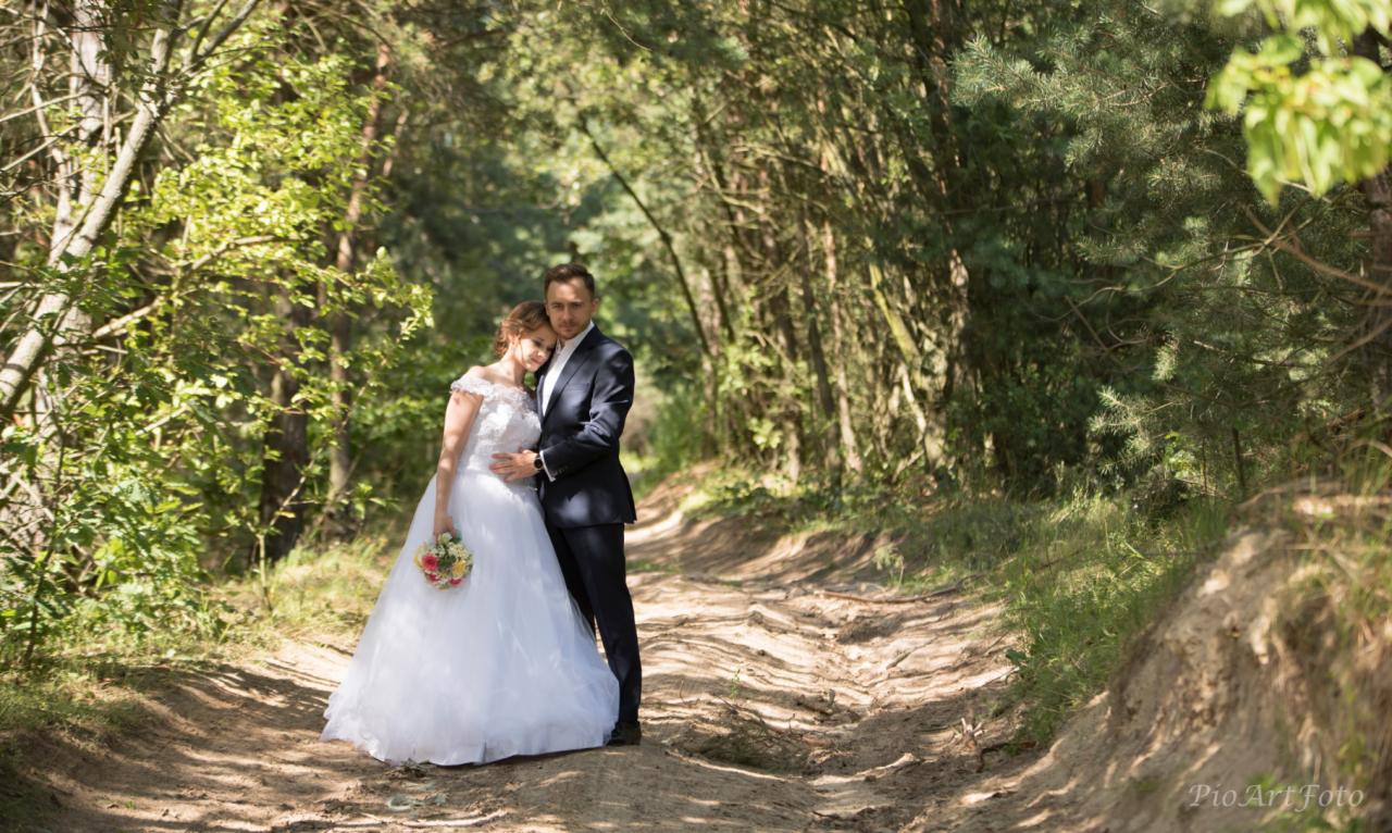 Para młoda w leśnej scenerii: Panna młoda w białej sukni z bukietem, pan młody w granatowym garniturze, stoją przytuleni na piaszczystej drodze, otoczeni drzewami.