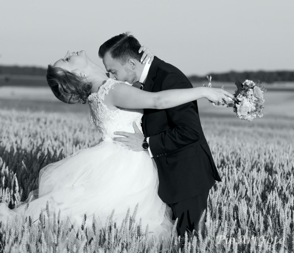 Czarno-białe zdjęcie: Panna Młoda w koronkowej sukni odchyla się, gdy Pan Młody w garniturze ją obejmuje na polu pszenicy.