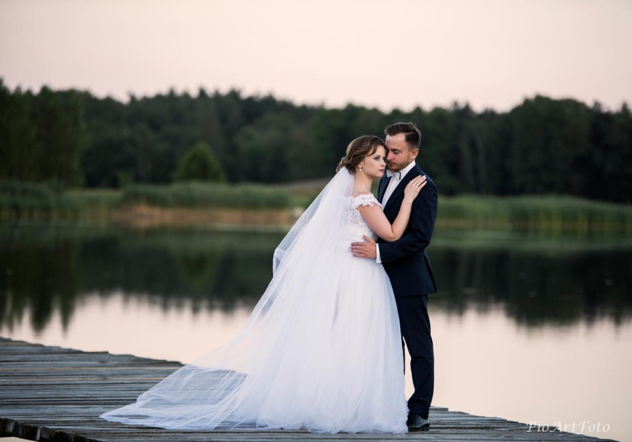 Para młoda w objęciach na drewnianym pomoście nad jeziorem, ona w koronkowej sukni ślubnej z długim welonem, on w granatowym garniturze, w tle las i zachodzące słońce.