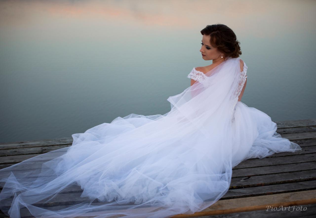 Panna młoda w białej sukni ślubnej z długim trenem siedzi na drewnianym pomoście nad wodą, spoglądając w bok.