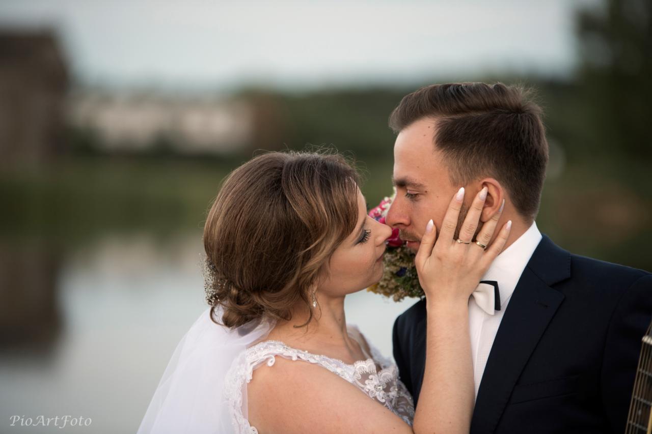 Panna młoda w białej sukni z koronkowymi zdobieniami i welonem dotyka dłonią policzka pana młodego w granatowym garniturze i białej muszce, zbliżając się do pocałunku; w tle rozmyty krajobraz.