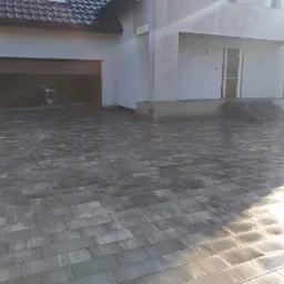 Ułożona szara kostka brukowa prowadząca do domu z otwartym garażem, widoczne schody wejściowe i fragment niedokończonego trawnika.