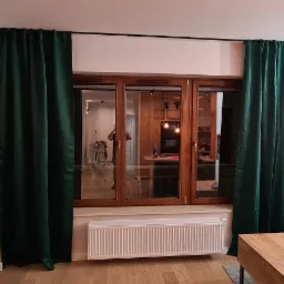 Salon z drewnianą stolarką okienną, zielonymi, aksamitnymi zasłonami, brązową sofą i drewnianym stolikiem kawowym z laptopem.