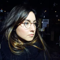 Portret kobiety w okularach o okrągłych oprawkach, z ciemnymi włosami z blond pasemkami, w ciemnym pomieszczeniu z dekoracjami w kształcie gwiazd