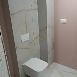 Nowoczesna toaleta podwieszana w łazience wykończonej płytkami imitującymi marmur z beżowymi żyłami i pudrowo różową ścianą, widoczne białe włączniki światła.