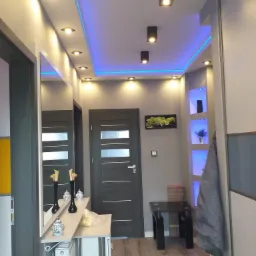 Nowoczesny przedpokój z szarą kolorystyką, oświetleniem LED podsufitowym, lustrem, oraz dekoracyjnymi elementami na półce, widok na drzwi wewnętrzne i wnękę z podświetlonymi półkami.