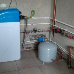 Domowa instalacja uzdatniania wody z niebiesko-białym zmiękczaczem, zbiornikiem wyrównawczym i rozbudowaną siecią rur miedzianych i plastikowych na tle ściany z fakturą.