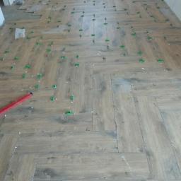 S.S.bud - Układanie podłogi z paneli imitujących drewno w jodełkę, z użyciem poziomicy i zielonych klipsów dystansowych.