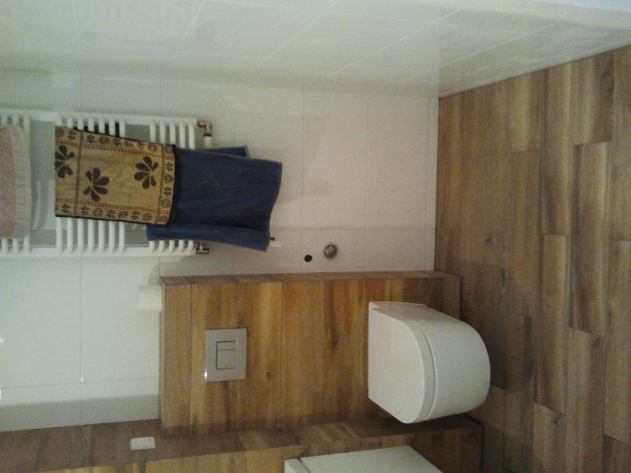 Nowoczesna łazienka z drewnianą okładziną ścienną, wiszącą toaletą i grzejnikiem drabinkowym z ręcznikami w beżowe kwiaty.