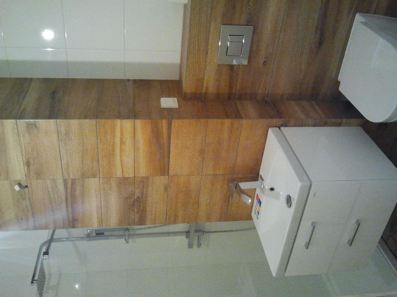 Nowoczesna łazienka z drewnopodobnymi płytkami na ścianach, białą armaturą i kabiną prysznicową z deszczownicą.