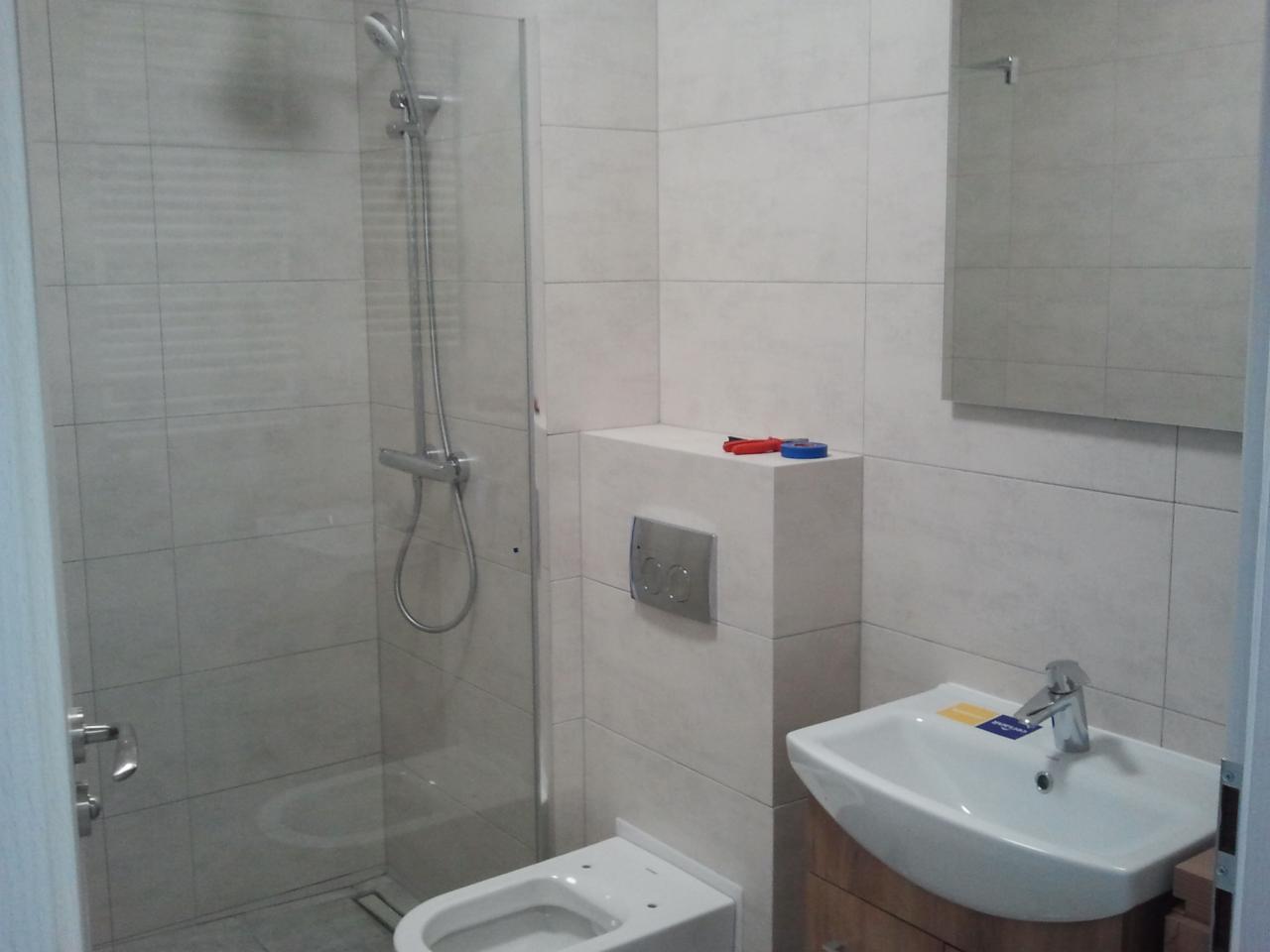 Nowoczesna łazienka z kabiną prysznicową typu walk-in, podwieszaną toaletą, umywalką nablatową na drewnianej szafce i szarymi płytkami. Na zabudowie toalety leżą czerwone i niebieskie narzędzia.