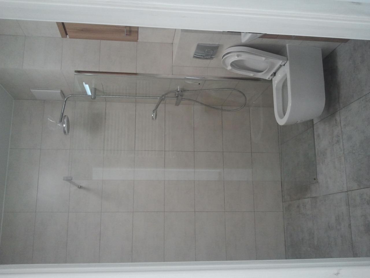 Nowoczesna łazienka z kabiną prysznicową typu walk-in, wiszącą toaletą z otwartą klapą i szarymi płytkami w dwóch odcieniach.