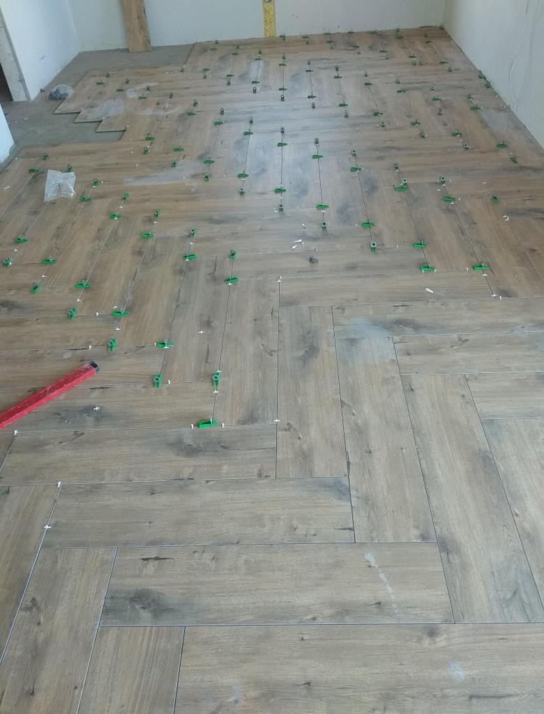 Układanie podłogi z paneli imitujących drewno w jodełkę, z użyciem poziomicy i zielonych klipsów dystansowych.