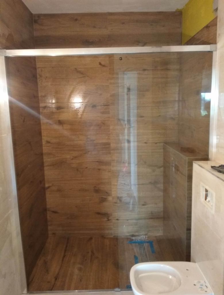 Nowoczesna kabina prysznicowa z drewnopodobnymi płytkami na ścianach i podłodze, przesuwne szklane drzwi z aluminiową ramą, widoczna miska WC w trakcie montażu.