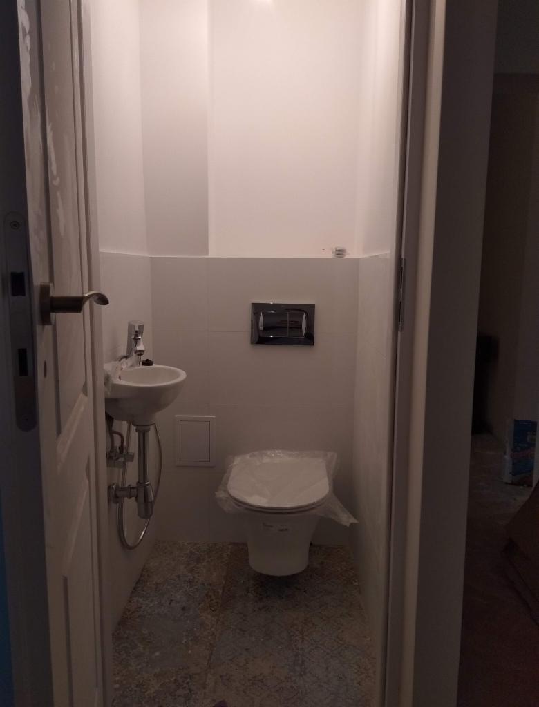 Mała łazienka z umywalką nablatową i wiszącą toaletą, zabezpieczoną folią ochronną, widok zza uchylonych drzwi.