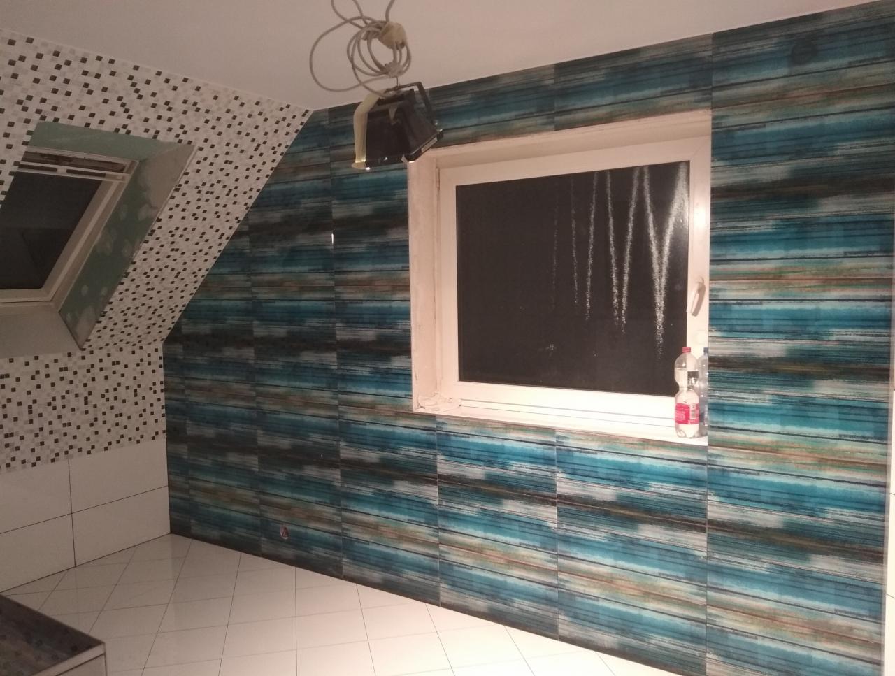 Łazienka w trakcie wykańczania: ściany wyłożone płytkami w odcieniach błękitu i brązu, mozaika przy oknie dachowym, białe płytki podłogowe, lampa robocza wisząca na kablu.