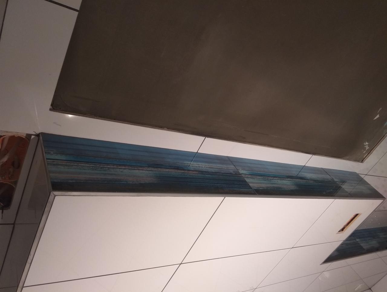 Niedokończona łazienka z białymi płytkami, dekoracyjnym pasem płytek z motywem morskim i surowym sufitem przygotowanym do malowania.