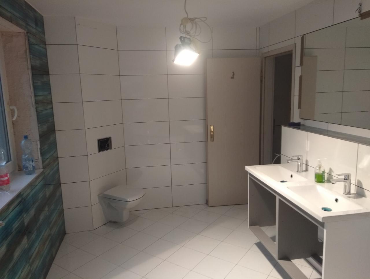 Jasna łazienka w trakcie wykańczania: białe płytki na ścianach i podłodze, podwieszana toaleta, biała umywalka z dwiema bateriami, lustro, okno z nieukończonym wykończeniem. Na oknie butelki.