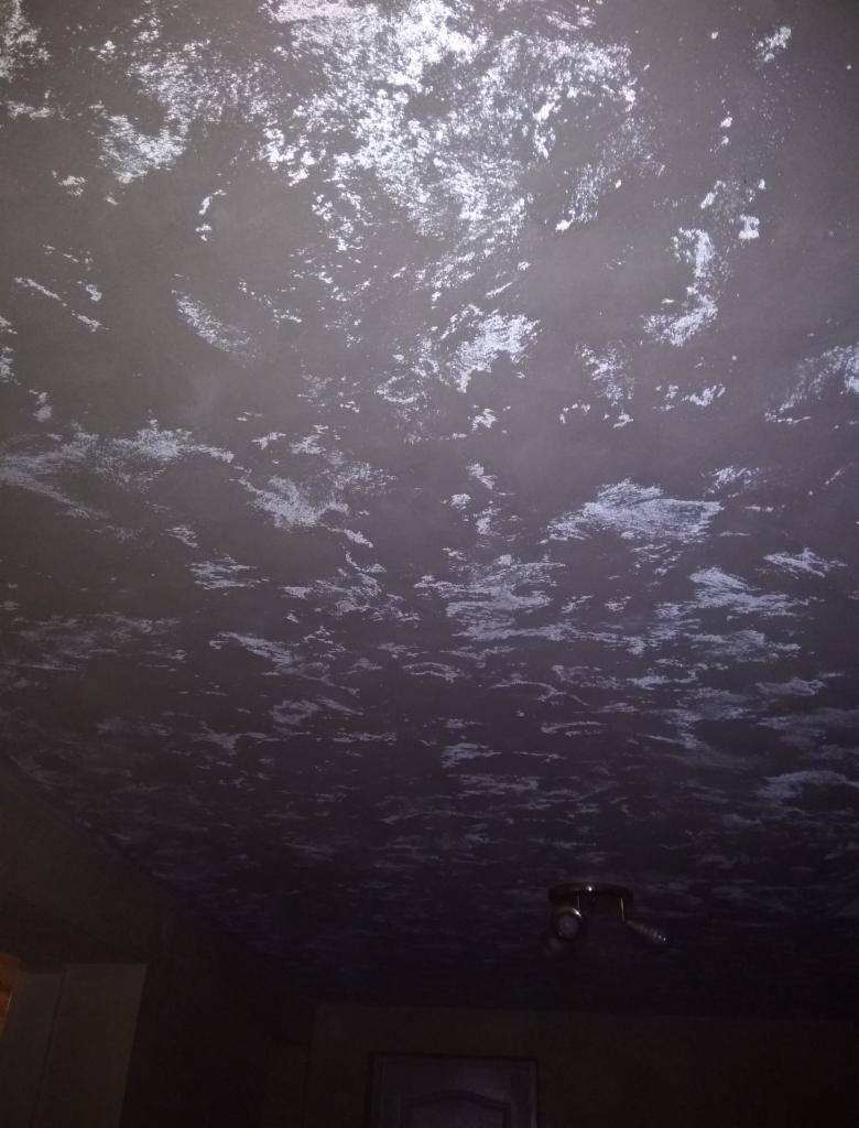 Sufit w ciemnym odcieniu szarości z nieregularnymi, jasnymi smugami, przypominającymi efekt dekoracyjny lub niedokładne położenie gładzi, widoczny fragment lampy sufitowej i drzwi.
