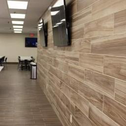 ORKAN Paweł Warpas - Ściana wyłożona podłużnymi płytkami imitującymi drewno, z dwoma zamontowanymi telewizorami LCD. Podłoga wyłożona płytkami w podobnym odcieniu. W oddali widoczne stoliki i krzesła.