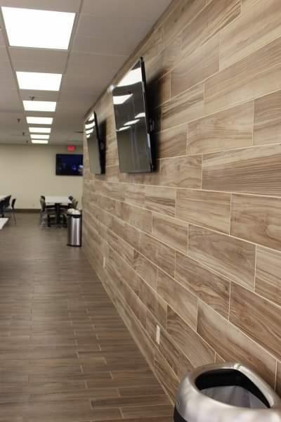 Ściana wyłożona podłużnymi płytkami imitującymi drewno, z dwoma zamontowanymi telewizorami LCD. Podłoga wyłożona płytkami w podobnym odcieniu. W oddali widoczne stoliki i krzesła.