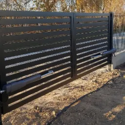 Nowoczesna, czarna brama przesuwna z automatyką, widoczne siłowniki i skrzynka sterująca, teren wokół bramy przygotowany pod dalsze prace wykończeniowe.